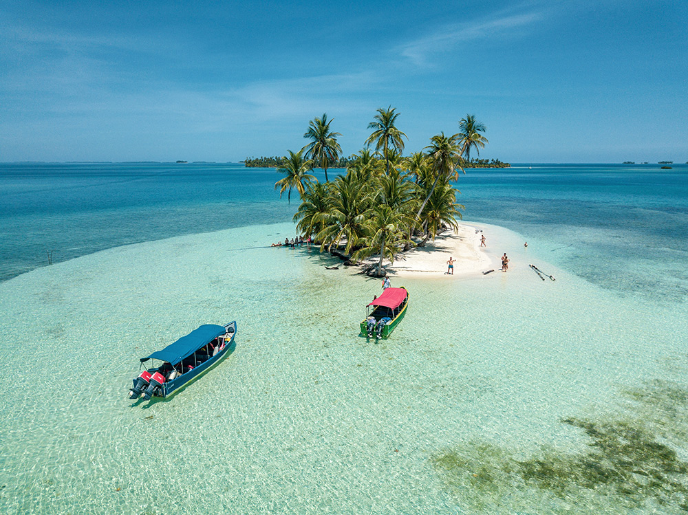 Aerial view of a small palm-covered San Blas island with two boats anchored in shallow turquoise water